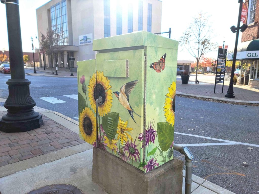 A photograph of a utility box covered with a colorful illustration of sunflowers, birds, and butterflies